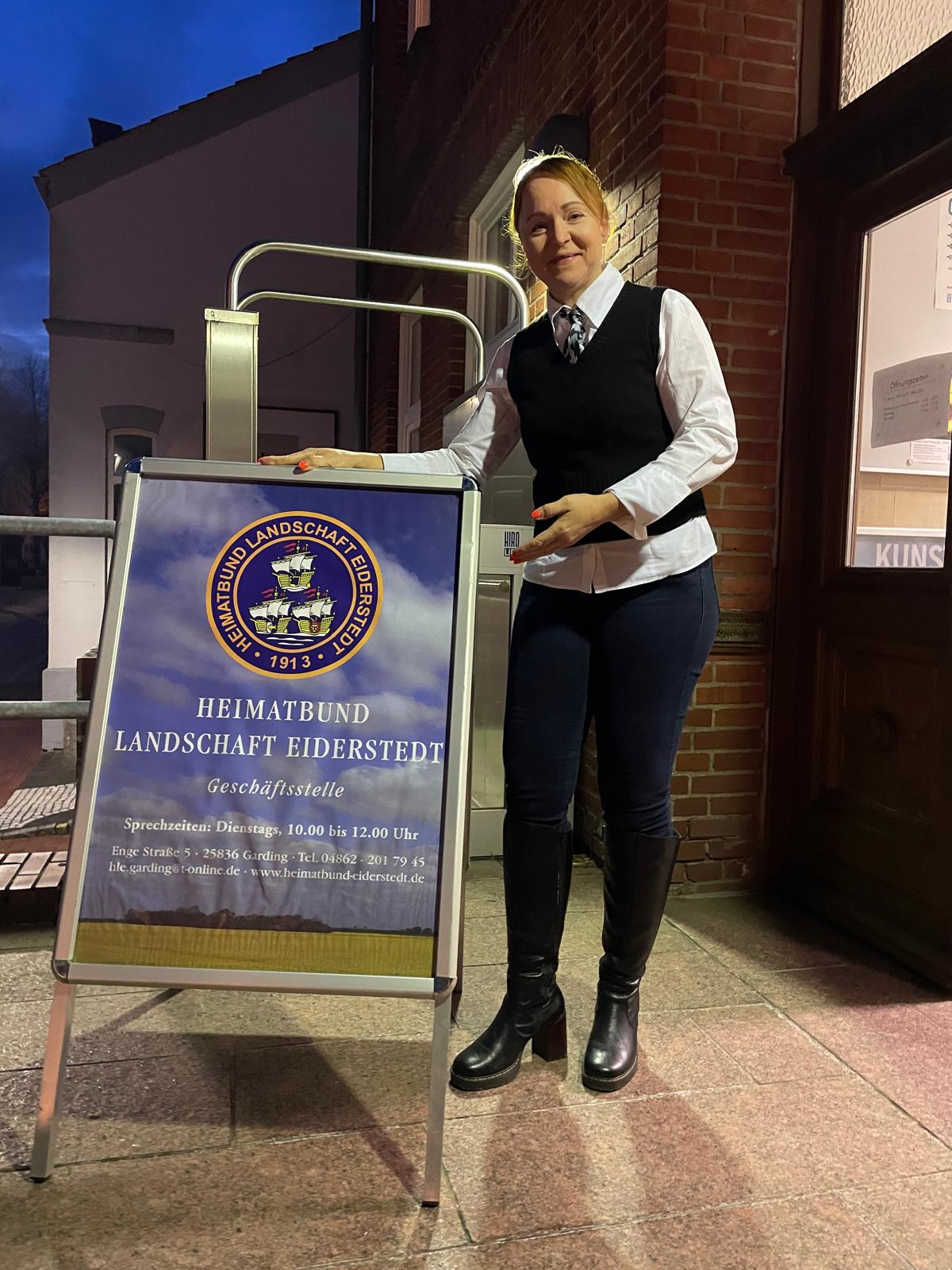 Isabella L&ouml;w steht am Aufsteller des Heimatbunds vor dem Kulturhaus/Stallerhaus in Garding